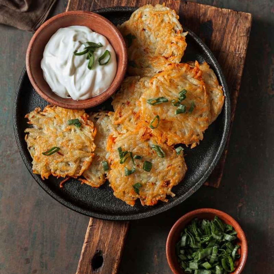 Crispy golden potato pancakes garnished with scallions, served on a black skillet with sour cream and chopped green onions.