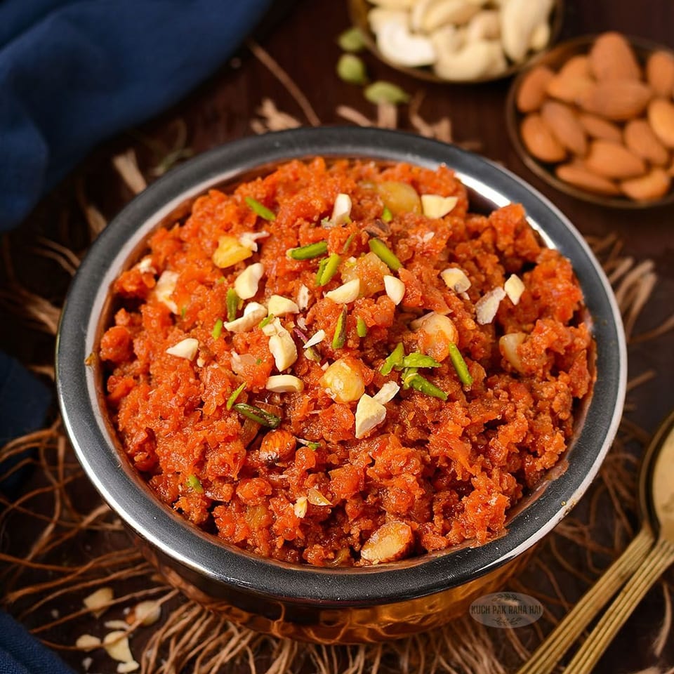 Bowl of vibrant orange Indian dessert garnished with chopped cashews and pistachios, surrounded by bowls of almonds and cashews.