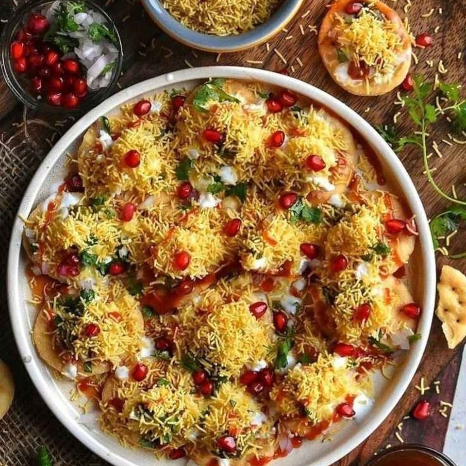 Large platter of Indian chaat topped with sev, pomegranate seeds, chutneys, yogurt, and cilantro, surrounded by bowls of garnishes.